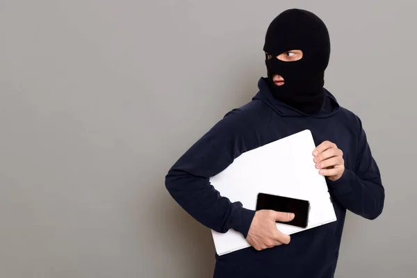 Man steals laptop and phone, runs away and turns back, being of persecution, wears robber mask and black turtleneck, advertising space, isolated over gray background.