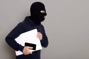 Çalıntı bir laptop, bir kadın çantası ve bir cüzdan ile kaçan bir suçlunun yan görüntüsü, soyguncu siyah bir kapüşonlu giyiyor, yüzü gizlenmiş, kopyalama alanı, gri arka planda izole edilmiş.