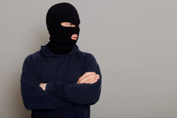 一个戴着强盗面具 头戴黑色帽衫 双手交叉放在胸前的罪犯站在那里 向旁边看去 抄袭广告空间 用灰色背景隔开 — 图库照片