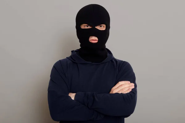 自信的犯罪男子 头戴黑色帽衫 戴着强盗面具 站在灰蒙蒙的背景上 看着摄像机 双手交叉放在胸前 摆出一副孤立无援的样子 — 图库照片