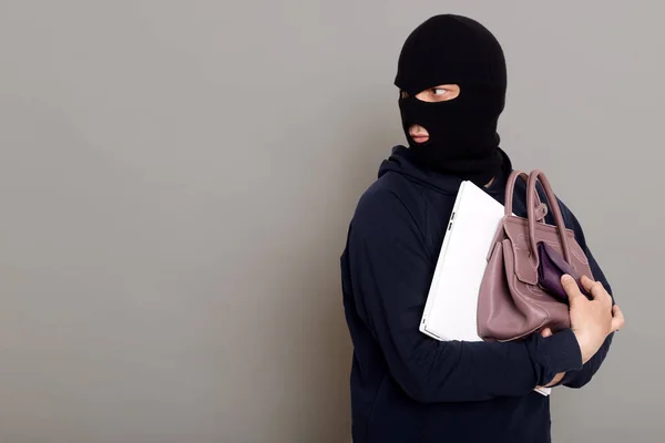 A young thief male dressed in a black hoodie with a disguised face runs away after stealing with other people\'s belongings, bag, laptop, wallet. Look around, be afraid of being caught, copy-space.