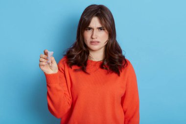 Genç ve çekici esmer kadın, gündelik giysiler giymiş, elinde regl kupası olan, hayal kırıklığına uğramış, üzgün, böyle bir alternatiften hoşlanmayan, mavi arka planda soyutlanmış bir kadın..