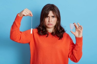 Koyu renk saçlı, turuncu kazaklı, elinde tampon ve fincanla poz veren, üzgün, ağlamak üzere olan, mavi arka planda soyutlanmış genç bir kadın..