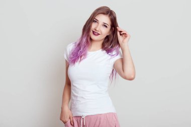 Parlak dudaklı ve leylak saçlı çekici genç bir kadın ayakta kameraya bakıyor, saçlarına dokunuyor, olumlu duygularını ifade ediyor, gri arka planda izole edilmiş bir şekilde poz veriyor..