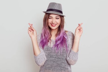 Tişört ve şapka giyen mutlu genç bir bayan, iyi bir şeyler umut ediyor, gözlerini kapalı tutuyor ve beyaz arka planda izole edilmiş, dişlek bir gülüşü var..