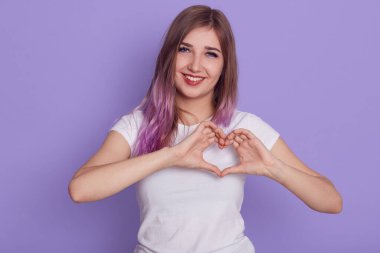 Good looking female with purple hair looking at camera with toothy smile, showing love symbol, making heart gesture with fingers, expressing positive, isolated over purple background. clipart