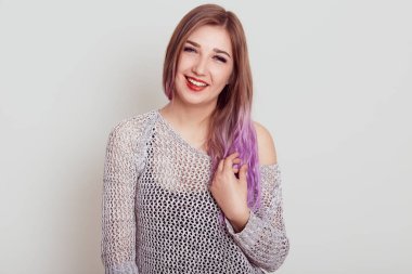 Gri gömlekli gülümseyen kadın kameraya bakıyor ve leylak saçlarına dokunuyor, olumlu duygularını ifade ediyor, mutlu görünüyor, gri arka planda izole edilmiş..