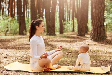 Küçük bebeğiyle ormanda yoga ve meditasyon yapan sportif bir kadın, güneşli bir günde parkta dinlenen siyah saçlı ve at kuyruklu beyaz elbiseli çekici bir anne..