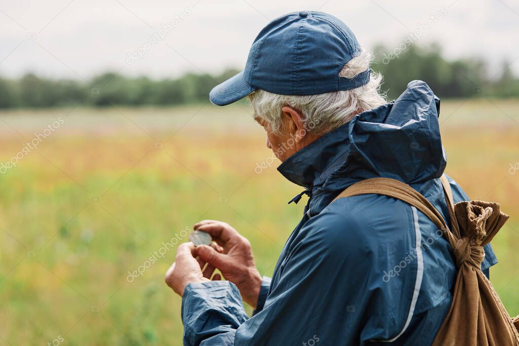 Gray haired man in jacket and cap treasure hunter looking at his finding, holding old coin under ground when searching in meadow or field.
