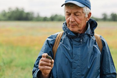 Middle aged male wearing blue jacket holding old coin found in meadow, looking attentively at his finding, numismatist posing outdoors with historical artifact.