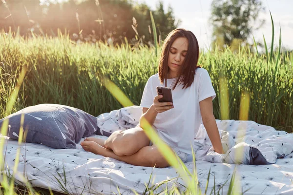 穿着白色休闲式T恤的黑发女人坐在草地上 用智能手机 上网或查看社交网站的形象 有着沉着的表情 — 图库照片