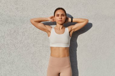 Ciddi koyu renk saçlı kadın portresi bej tozlu ve beyaz bluzlu gri arka planda soyutlanmış, kollarını kaldırmış, sağlıklı bir yaşam tarzı var..