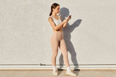 Beyaz bluzlu ve bej tozluklu esmer bir kadının tam boy profil portresi. Elinde akıllı bir telefon, internet üzerinden iletişim kurmak için cep telefonu kullanıyor..