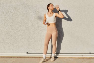 Beyaz bluzlu, bej tozluklu, spor egzersizleri yaptıktan sonra su içen zayıf kadın portresi açık havada çalışırken susar..