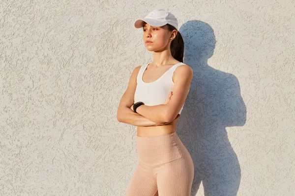 Outdoor shot of confident girl wearing visor cap, white top and beige leggins, folded hands, having serious facial expression, posing against gray wall outdoor.