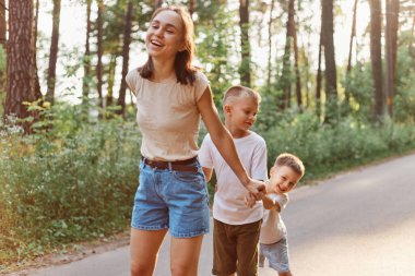 Doğada oynayan iki küçük oğlu olan bir anne, tişörtlü koyu renk saçlı bir kadın ve güneşli bir yaz gününde çocuklarıyla vakit geçirmek, aile, çocukluk..