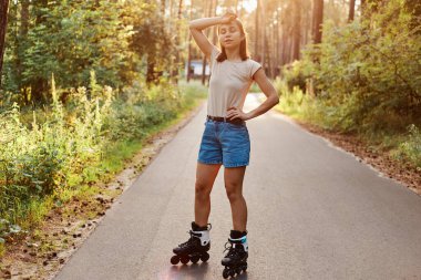 Yaz parkında yolda duran yorgun kadın, patenden bitap düşmüş, dinlenmek için duruyor, elini alnına koyuyor, kapalı gözlerle dikiliyor, günlük giysiler giyiyor..
