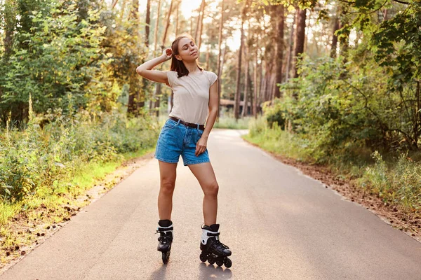 在公园的沥青路面上 身穿米色T恤和牛仔裤衬衫的健康的年轻的黑发女子骑在溜冰鞋上 享受着积极的生活 紧闭双眼 — 图库照片