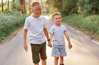 Two little boys wearing white t shirts and shorts going together and holding hands in summer park, brothers walking outdoor, expressing positive emotions. clipart