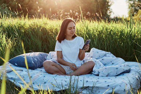 在草地上 穿着白色T恤 双腿交叉坐在柔软的床上 用智能手机 手持手机 查看社交网络的年轻成年黑发女性的画像 — 图库照片