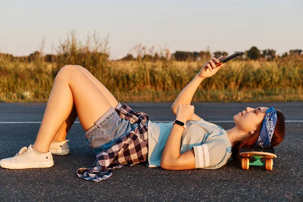 Dark haired young beautiful lady lay down on the asphalt road outdoor with hand on skateboard, using a mobile phone to take selfie or cheking social networks.
