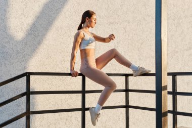 Beyaz bluzlu ve bej tozluklu bir kadın, korkuluklara oturmuş akıllı saatine bakıyor, spor yapıyor..