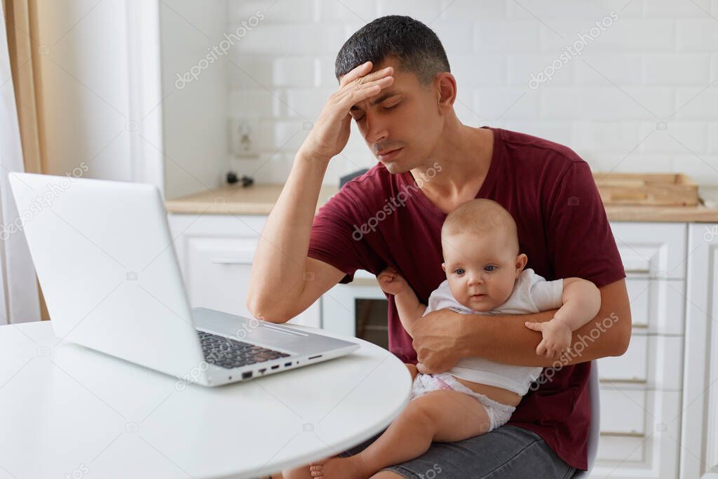 Indoor image of a man working from home with the baby on his laps, tired young adult father with small toddler baby working in kitchen, being exhausted, lack of sleep.