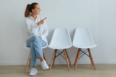 Beyaz gömlek ve kot pantolon giyen genç bir kadın sandalyede oturuyor ve işaret parmağıyla bir tarafa bakıyor. Koyu renk saçlı ve at kuyruklu. Keyfi yerinde..