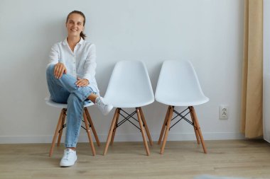 Arkadaşça gülümseyen iyimser bir kadın. Kapalı alanda, ışık duvarına yaslanmış, mutlu ve kendinden emin bir ifadeyle kameraya bakıyor. Beyaz gömlek ve kot pantolon giyiyor..