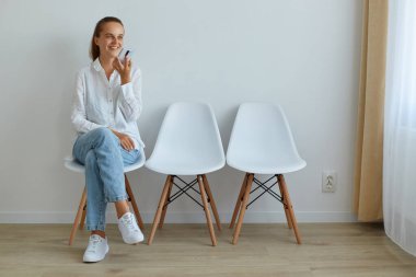 Sandalyeye oturmuş gülümseyen genç bir kadının portresi, elinde akıllı bir telefon, cep telefonunda ses asistanı istemek, görev vermek, mesaj kaydetmek, olumlu duygular ifade etmek..