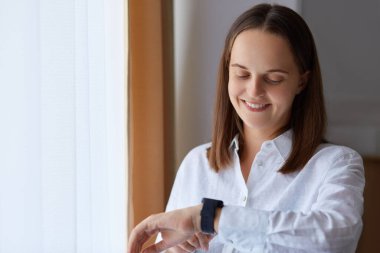 Beyaz gömlek giyen, gülümseyen genç bir kızın portresi mutlu bir ifadeyle kol saatine bakıyor, pencerenin yanındaki aydınlık odada poz veriyor, zevk alarak ukalalık yapıyor..