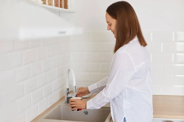 stock image Side view portrait of dark haired woman wearing white shirt standing near kitchen set, pouring water from faucet in the kitchen, washing mug, posing indoor in light room.