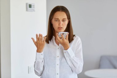 Kafası karışmış beyaz gömlekli bir kadının portresi evde elinde akıllı bir telefonla poz veriyor, kolunu kaldırıyor, cihazın neden çalışmadığını anlamıyor..