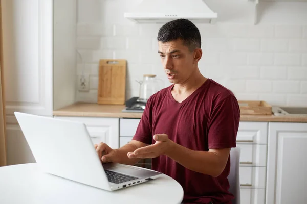 白いテーブルのキッチンに座っている若い大人の男性のフリーランスの屋内ショット コンピュータ上で作業 仕事上の問題を抱えています 困惑式のPc画面を見て — ストック写真