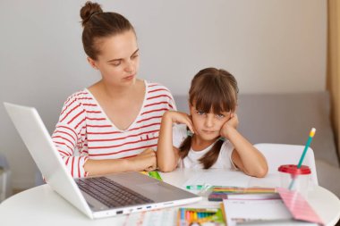 Yoğunlaşmış kadın, kitap ve dizüstü bilgisayarla masada kızıyla oturuyor, ödev yapıyor ya da internetten ders alıyor, anne çocuğuna yardım ediyor, uzaktan eğitim alıyor..