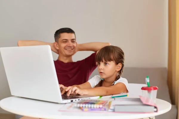Happy positive man sitting with raised looking at his daughter who sitting at table and looks at lap top display with scared expression, family posing at home.