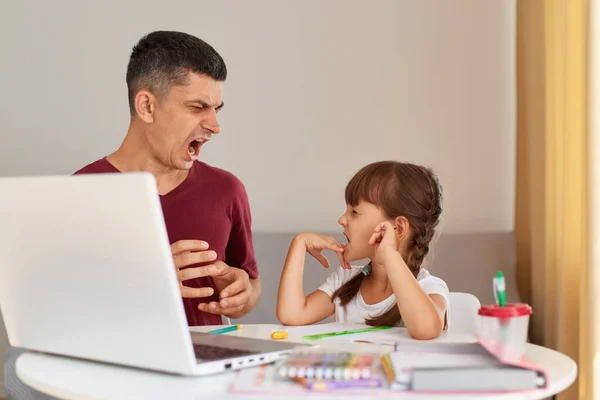 Indoor shot of angry aggressive father screaming at his daughter while helping her to do homework, family posing in room at home while sitting at table.