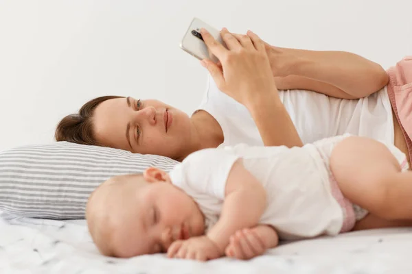 睡眠不足的女儿穿着白色衣服躺在母亲身边 女人醒来后用智能手机查看电子邮件或社交网络的形象 — 图库照片