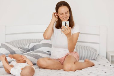 Beyaz tişörtlü ve şortlu bir kadının elinde akıllı bir telefon tutarken, cihaz ekranına bakarken, yatağında bez bezli küçük bir bebekle otururken..