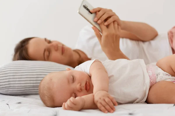 睡梦中穿着白色T恤的婴儿 躺在女儿身边拿着手机的母亲 使用智能手机上网或打字的妇女的室内照片 — 图库照片