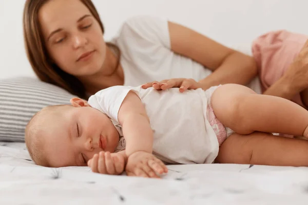 若いです大人の女性と暗い髪はベッドの中で赤ちゃんと横たわっています 娘を見て 彼女は眠っているかどうかを見て 女性は白いカジュアルなTシャツを着て 幸せな母親 — ストック写真