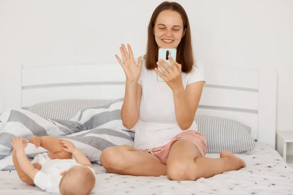 快乐的女人穿着白色T恤和短裤 有视频通话或直播 挥手对着摄像机笑着 抱着小孩坐在床上摆姿势 — 图库照片