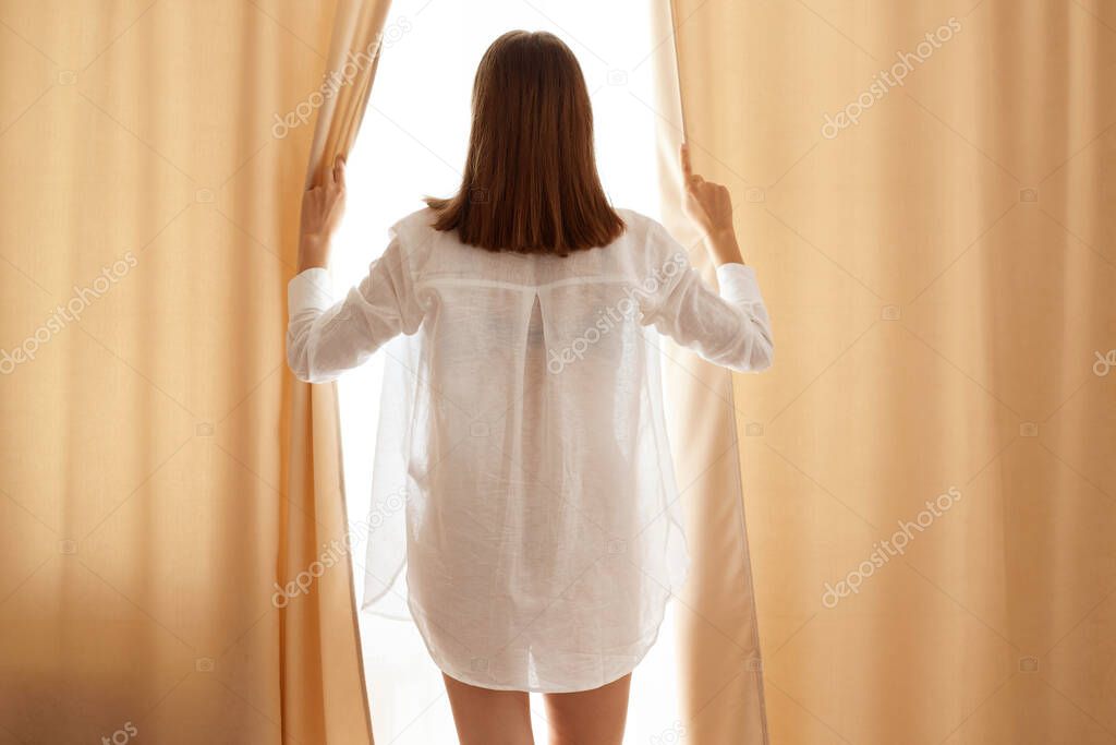 Back side portrait of dark haired female wearing cotton white shirt open curtains, standing near window, woman with beautiful perfect body posing backwards at home.