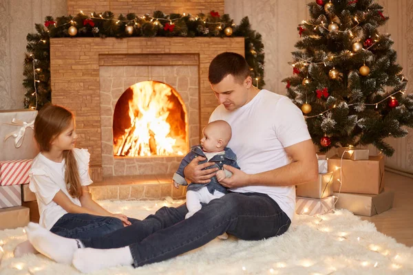 在室内拍摄的照片中 年轻的白人父亲和两个女儿坐在壁炉边和圣诞树旁的地板上 一起庆祝寒假 — 图库照片