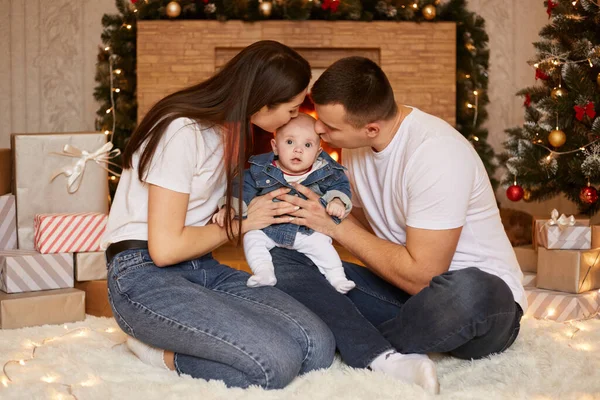 年轻家庭亲吻小女儿的肖像 坐在圣诞树旁和壁炉旁的地板上 在家里共度和庆祝寒假 — 图库照片
