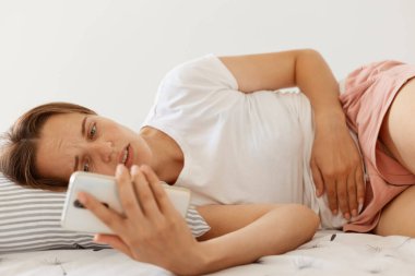 Suratı asık, beyaz tişörtlü hasta bir kadın yatakta uzanmış, elinde akıllı bir telefonla, korkunç bir mide ağrısından muzdarip bir doktoru aramak zorunda..