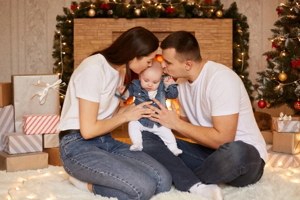 屋内ショットの若いです幸せな家族のポーズでクリスマスお祝いの部屋中に床に座って一緒にキス彼らの小さなかわいい赤ちゃんの娘 陽気なクリスマスと幸せな新年 — ストック写真