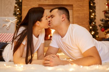 Güzel esmer, beyaz tenli, romantik bir çift yeni yıl arifesini kutluyor, beyaz yumuşak halının üzerinde çelenk ışıklarıyla yerde öpüşüyorlar..