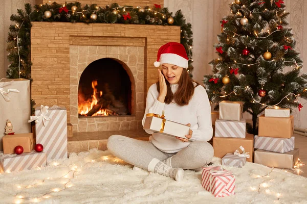 白いセーターとサンタクラスの帽子を身に着けている驚きの女性交差脚と開いているクリスマスギフトボックスに座って 頬に手のひらを保ち 冬の休日のために存在することに驚いています — ストック写真