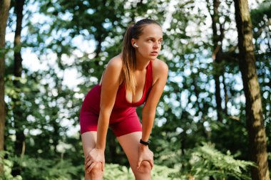 Kırmızı atletik takım elbiseli yorgun ama kararlı bir kadın ellerini dizlerinin üzerine uzatarak ormanda koşuya ara veriyor. Kablosuz kulaklık takıyor..